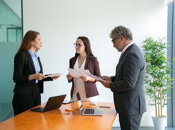 Successful coworkers talking, holding papers and working together. Professional confident office employees standing at table with laptops in conference room. Business, teamwork and management concept